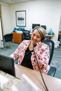 A woman is sitting at a desk in an office, smiling while talking on the phone