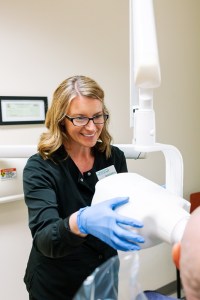 A dental professional, wearing blue gloves and a black top, is assisting a patient in a dental office. 