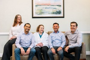 A photo of a group of five dental professionals is sitting together on a couch in a professional or office setting